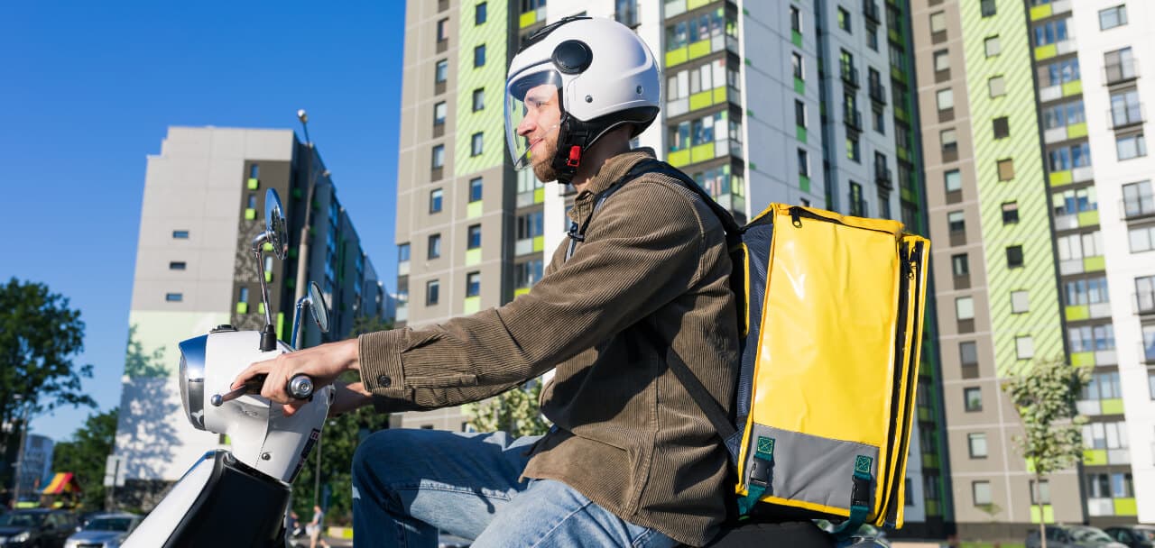 Mensajero en moto con caja de reparto circulando por una vía urbana (imagen de referencia)